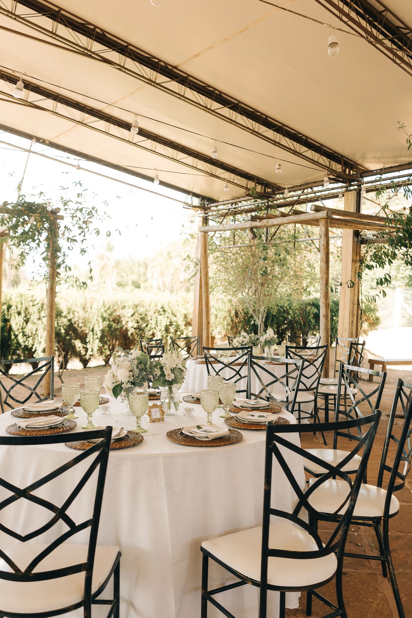 Tables rondes blanches sous une pergola fleurie, verres verts et chaises en fer forgé
